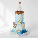 A two-tiered butter cream cake with a nautical theme, featuring sailboats, waves, and themed decorations in blue shades, celebrating a birthday with 'Happy Birthday' text and a age '2' displayed.