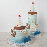 A beautifully decorated butter cream cake with a nautical theme, featuring edible sailboats, anchors, and ocean waves on a blue background.