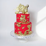 A two-tiered fondant cake decorated with gold dragon designs and Chinese characters, primarily in red and gold colors, displayed on a white cake stand.